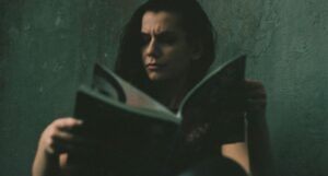 fair-skinned person with long hair reading a book against a greenish background